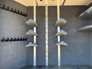 Interior of a Cimarron 4 Horse Trainer trailer featuring carpeted walls, six angled shelves mounted vertically in the center, black hooks on the left wall, and two metal shelves attached to the right wall. -Stillwater Trailer Sales