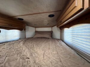 A neatly made bed with a brown quilt and pillow is situated in a compact, wood-paneled LQ space—ideal for a 3 Horse Trails West trailer—with cabinets above and windows on both sides covered by light-colored blinds. -Stillwater Trailer Sales