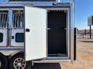 A silver Cimarron horse trailer with an open tack room door reveals empty saddle racks inside. This 4 Horse Trainer trailer is parked on gravel near a road, with a TRAILER SALES sign visible in the background. -Stillwater Trailer Sales