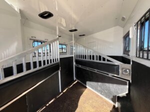 Interior of a Trails West horse trailer with white walls, metal dividers, barred windows, and padded black panels along the sides. Sunlight enters through the windows, illuminating the partitioned stalls of this 3 Horse LQ. -Stillwater Trailer Sales