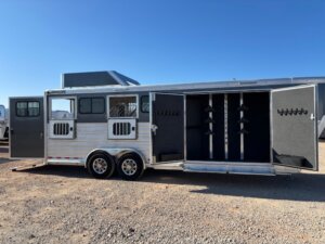 A silver Cimarron gooseneck 4 Horse Trainer trailer with two open tack room doors showing saddle racks and bridle hooks, plus side windows with bars; parked on gravel under a clear blue sky. -Stillwater Trailer Sales