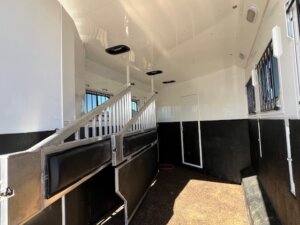 Interior of a Trails West 3 Horse LQ trailer features partitioned stalls, metal bars, padded dividers, black lower walls, white upper walls, and multiple windows with bars for ventilation. The floor has a textured surface for added safety. -Stillwater Trailer Sales