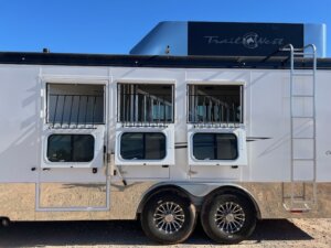 Side view of a Trails West 3 Horse LQ trailer with three open barred windows, matching lower access doors, two shiny wheels, a metal ladder on the right, and logo on top against a clear blue sky. -Stillwater Trailer Sales