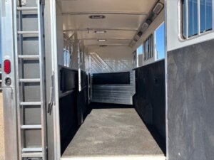 The image shows the inside of a Cimarron 4 Horse Trainer trailer with rubber flooring, padded side walls, a dividing gate, and a ladder mounted on the left near the door. Natural light enters through the side windows. -Stillwater Trailer Sales