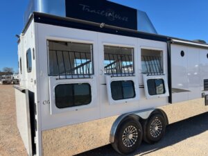 A white Trails West 3 Horse LQ horse trailer with three side windows featuring metal bars and two axles with shiny wheels is parked outdoors on a sunny day. The brand name is visible on the upper section. -Stillwater Trailer Sales