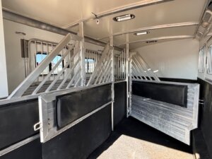 Interior of a Cimarron 4 Horse Trainer trailer showing divided stalls with metal partitions, padded panels, and grated windows. The space appears clean and well-lit with a non-slip floor. -Stillwater Trailer Sales