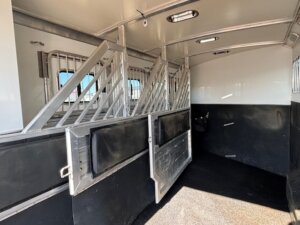 Interior of a Cimarron 4 Horse Trainer trailer showing metal dividers, padded side panels, windows with bars, and open stall doors. The black and white walls and non-slip floor ensure comfort and safety for your horses. -Stillwater Trailer Sales