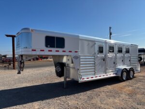 A silver Elite 4 Horse gooseneck trailer with three windows and three doors on the side, parked on gravel near a road under a clear blue sky. -Stillwater Trailer Sales
