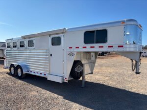 An Elite 4 Horse silver gooseneck trailer with three windows and dual axles is parked on gravel under a clear blue sky. The trailer features a ribbed side and a spare tire mounted near the front. -Stillwater Trailer Sales