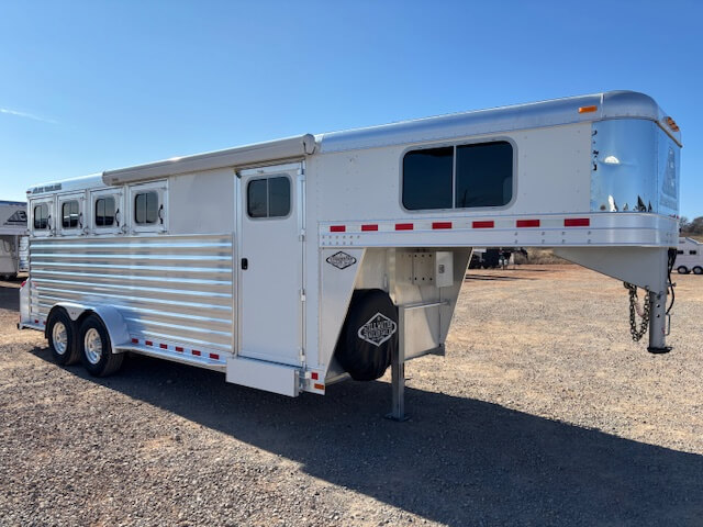 An Elite 4 Horse silver gooseneck trailer with three windows and double axles is parked on a gravel lot under a clear blue sky. The trailer features a side door, spare tire, and red reflective tape along the sides. Horse 16400 model. -Stillwater Trailer Sales
