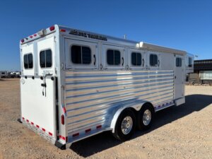 An Elite 4 Horse silver and white trailer with four windows and two axles is parked on a gravel lot under a clear blue sky. -Stillwater Trailer Sales
