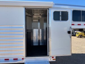 An open door on the side of a white Elite 4 Horse trailer reveals a clean, empty interior with metal walls. In the background, a yellow truck and another trailer are visible. -Stillwater Trailer Sales