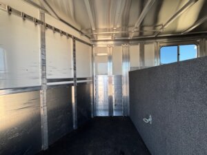 Interior of an empty Elite 4 Horse metal trailer with a partially carpeted divider, hooks along the upper left wall, a small window on the right, and sunlight streaming through the open door. Horse 16400. -Stillwater Trailer Sales