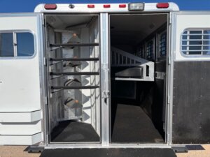An Elite 4 Horse trailer with its back doors open, showing multiple saddle racks on the left side and a walkway leading to individual horse stalls on the right. The clean and empty interior is ready for your horses. -Stillwater Trailer Sales