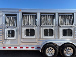 A close-up side view of an Elite 4 Horse trailer with four barred windows, lower ventilation doors, and two large wheels visible, parked under a clear blue sky. -Stillwater Trailer Sales