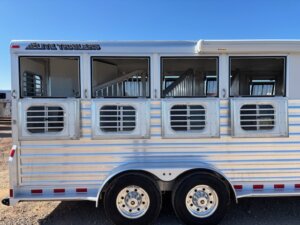 A silver Elite 4 horse trailer with three open windows showing interior stalls, double axles with black and white wheels, and the words ELITE TRAILERS on the upper left side, parked on gravel under a clear blue sky. Stock #16400. -Stillwater Trailer Sales