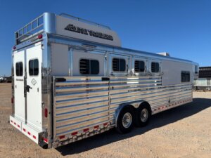 A large silver and white Elite 4 Horse trailer with four windows and dual axles is parked on gravel under a clear blue sky. The trailer features Elite Trailers signage above the roof and boasts a finished dressing room for added convenience. -Stillwater Trailer Sales