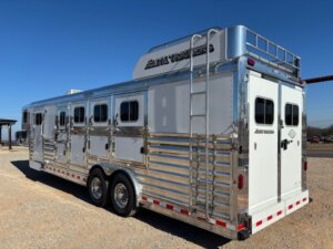 A large silver and white Elite 4 Horse trailer with multiple side doors, rear double doors, rooftop storage, ladder, and a finished dressing room, parked on gravel under a clear blue sky. -Stillwater Trailer Sales