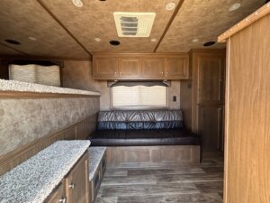 View of an RV interior with wood cabinets, a leather couch under a window with a closed blind, overhead storage, carpeted bench surfaces, wood-paneled flooring, and a ceiling vent—ideal for an Elite 4 Horse trailer’s finished dressing room. -Stillwater Trailer Sales