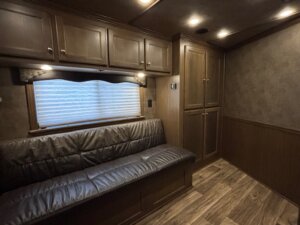 A finished dressing room with wood-paneled walls and flooring, featuring a built-in leather couch under a window with closed blinds, overhead cabinets, and tall storage cabinets on the right—perfect for any Elite 4 Horse trailer. Ceiling lights provide ample illumination. -Stillwater Trailer Sales