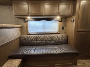A brown leather couch sits in the Finished Dressing Room of the Elite 4 Horse trailer, set against a wooden wall with overhead cabinets, under a window with a pleated shade and two small reading lights. Wood paneling and a speckled countertop complete the area. -Stillwater Trailer Sales
