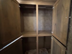 Wooden cabinet with four open doors, revealing empty shelves inside, reminiscent of storage found in a finished dressing room or elite 4 horse trailer. The interior has a brown finish and textured back panel, set against a neutral-colored wall. -Stillwater Trailer Sales