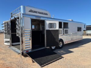 An Elite 4 Horse trailer with multiple windows and vents is parked on gravel. The side and rear doors are open, revealing a finished dressing room inside and a black ramp extended to the ground beneath the clear blue sky. -Stillwater Trailer Sales
