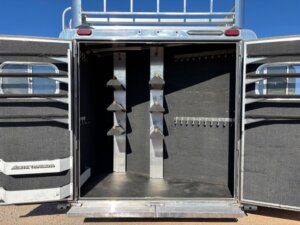 Open rear compartment of Elite 4 Horse Trailer with black interior, featuring three angled saddle racks on the left and multiple bridle hooks on the right, both doors open in sunlight, leading to a finished dressing room. -Stillwater Trailer Sales