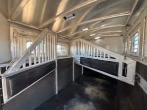 Interior view of an Elite 4 Horse trailer featuring padded dividers, rubber flooring, barred partitions, and side windows allowing natural light inside. Includes a finished dressing room for added convenience. -Stillwater Trailer Sales