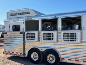 A silver Elite 4 Horse Trailer with barred windows and double axles is parked on a gravel lot beneath a clear blue sky. The trailer features Elite Trailers branding on the upper section and includes a finished dressing room. -Stillwater Trailer Sales