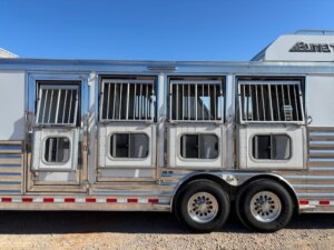 A close-up side view of an Elite 4 Horse Trailer with four barred windows and four small access doors, plus a finished dressing room, parked on gravel under a clear blue sky. -Stillwater Trailer Sales