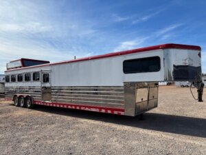 A large Sundowner silver and red 6 Horse gooseneck trailer with multiple windows and three axles is parked on a gravel lot under a blue sky. -Stillwater Trailer Sales