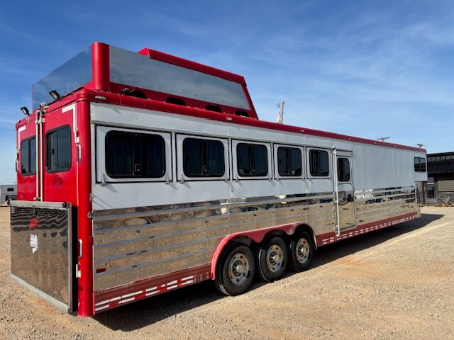 A large red and silver Sundowner horse trailer with six windows on the side is parked on a gravel lot under a clear blue sky. The spacious 6 horse trailer features triple axles and a raised observation deck or storage area on top. -Stillwater Trailer Sales