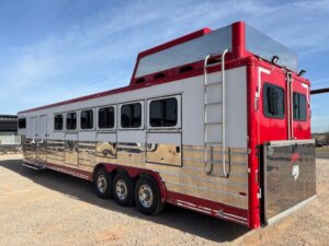 A large, red and silver Sundowner gooseneck horse trailer with multiple windows, three axles, a rear ladder, and reflective panels sits parked on gravel under a blue sky. Ideal as a spacious 6 horse trailer. -Stillwater Trailer Sales