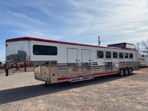 A large Sundowner horse trailer with triple axles, shiny metal sides, red roof and trim, several windows, and multi-compartment space for up to 6 horses is parked on a gravel lot under a partly cloudy sky. -Stillwater Trailer Sales
