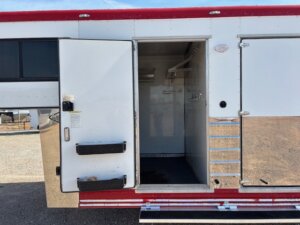 A white and red Sundowner horse trailer with an open side door reveals an empty, clean interior. The trailer features metal steps, a handle, and is parked on a gravel surface under a clear sky. -Stillwater Trailer Sales
