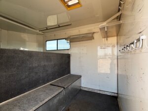 Interior of a Sundowner 6 Horse trailer with gray carpeted bench seating along one wall, a small window, metal shelves, and hooks mounted on a white wall, with natural light streaming through an overhead hatch. -Stillwater Trailer Sales