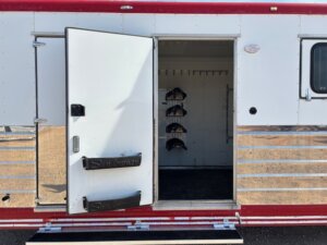 A Sundowner horse trailer with a white and red exterior is shown with its door open, revealing an interior area equipped with three saddle racks mounted on the wall. -Stillwater Trailer Sales