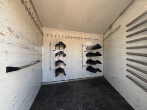 Interior of an empty white Sundowner horse trailer with multiple metal hooks on the walls, racks holding black wheel chocks, and a dark floor showing signs of wear. Ideal for use as a 6 horse transport setup. -Stillwater Trailer Sales