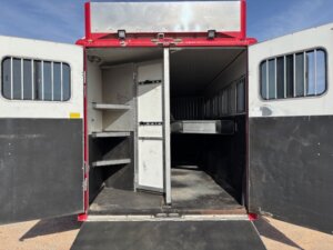 Open rear doors of a red-and-white Sundowner horse trailer reveal an empty, divided interior with metal bars, shelves on the left side, and a black ramp extending to the ground under a clear sky. Ideal for 6 horse transport. -Stillwater Trailer Sales