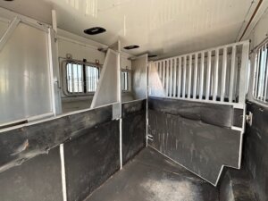The interior of this Sundowner 6 Horse Trailer features divided stalls, metal bars, and padded walls. Sunlight enters through barred windows along one side, highlighting the clean and well-maintained surfaces. -Stillwater Trailer Sales