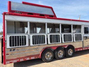 A Sundowner 6 Horse trailer in red and silver, featuring six barred windows, three axles, and reflective panels, is parked on gravel under a clear sky. -Stillwater Trailer Sales