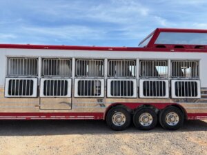 A large red and white Sundowner horse trailer with barred windows, three axles, and space for 6 horses is parked on dirt under a partly cloudy sky. -Stillwater Trailer Sales