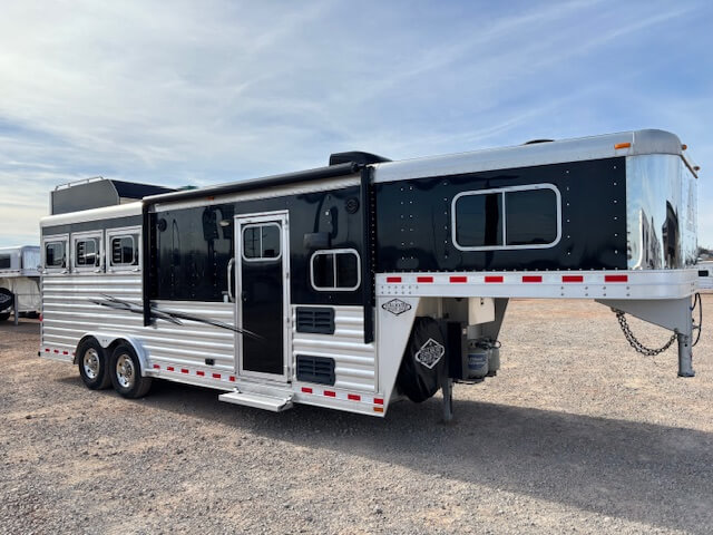 A large, silver and black Elite 3 Horse Trailer with Outback LQ living quarters is parked on gravel under a mostly clear sky. The trailer features multiple windows, a side door, and three axles. -Stillwater Trailer Sales