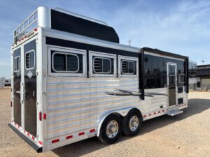 A silver and black Outback LQ Elite 3 Horse trailer with three side windows, double rear doors, two axles, and a small awning is parked on gravel under a blue sky. -Stillwater Trailer Sales