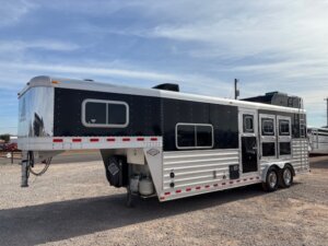 A black and silver Elite 3 Horse gooseneck trailer with three windows and stalls is parked on a gravel lot under a partly cloudy sky. -Stillwater Trailer Sales