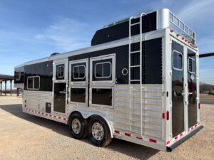 An Elite 3 Horse gooseneck trailer with black and silver accents, multiple windows, and compartments is parked on gravel. This horse trailer features dual axles, a ladder, roof storage, and rear double doors under a clear sky. -Stillwater Trailer Sales