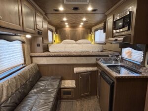 Interior of an Elite 3 Horse Outback LQ horse trailer featuring a kitchenette with sink and microwave, leather sofa, and raised bed area with steps. Wood cabinetry and windows with blinds are visible throughout. -Stillwater Trailer Sales
