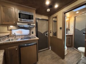 Interior of a compact Outback LQ horse trailer featuring a kitchenette with sink, small fridge, microwave, and cabinets; a doorway leads to a bathroom with toilet and sink; brown and beige color scheme throughout. -Stillwater Trailer Sales