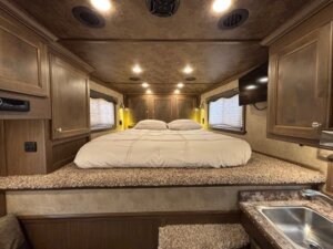A cozy bedroom in an Outback LQ horse trailer with a raised bed covered in beige bedding, wooden cabinets, two side windows with blinds, ceiling lights, and a wall-mounted TV on the right. A sink is partially visible below. -Stillwater Trailer Sales